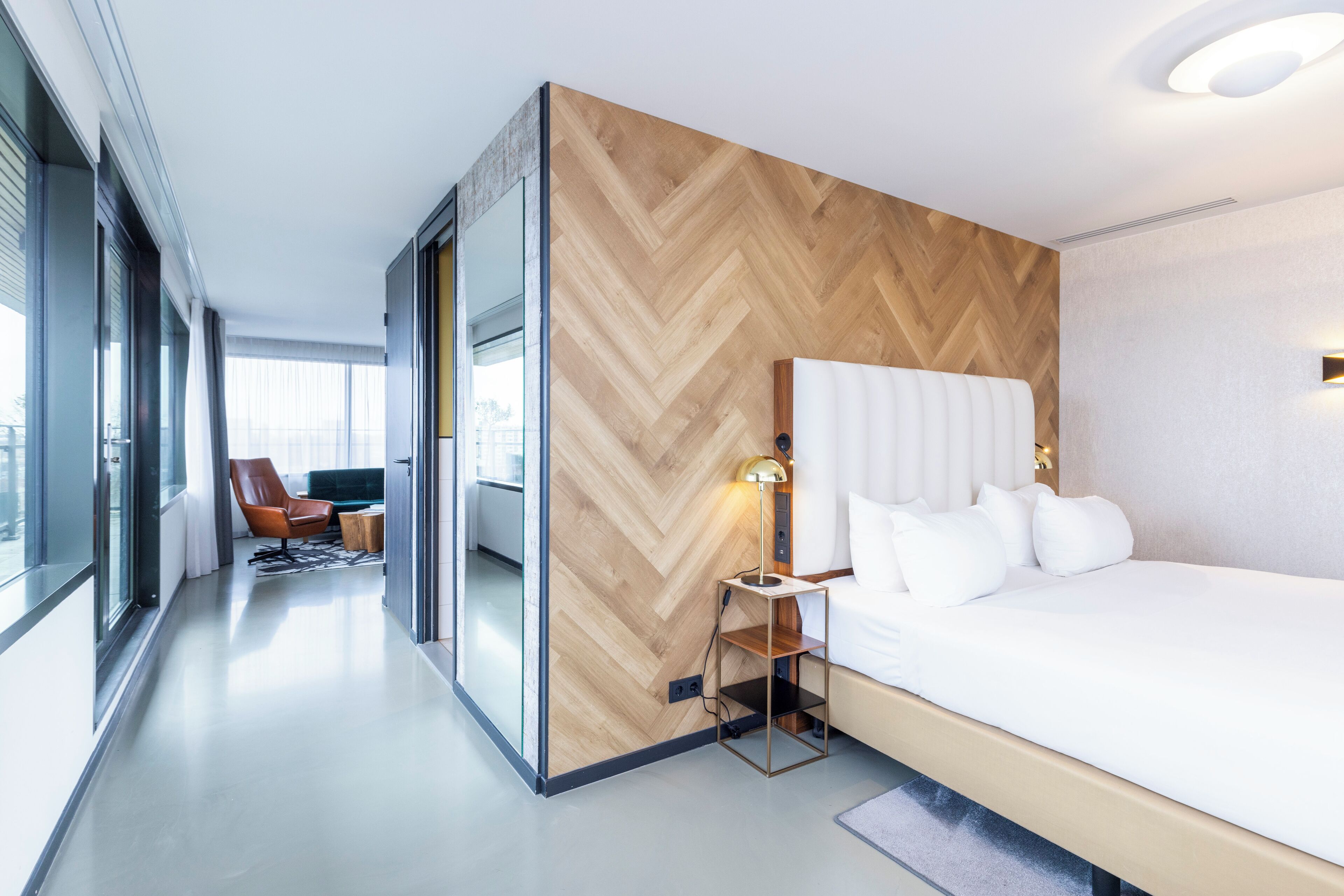 Una habitación de hotel moderna con una cama grande, una pared de madera con patrón de espiga, una mesita de noche pequeña con una lámpara, un sillón de cuero y vistas amplias a través de grandes ventanales.
