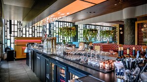 Zona de bar con barra, vasos, botellas y un área de estar con pared estampada y plantas.
