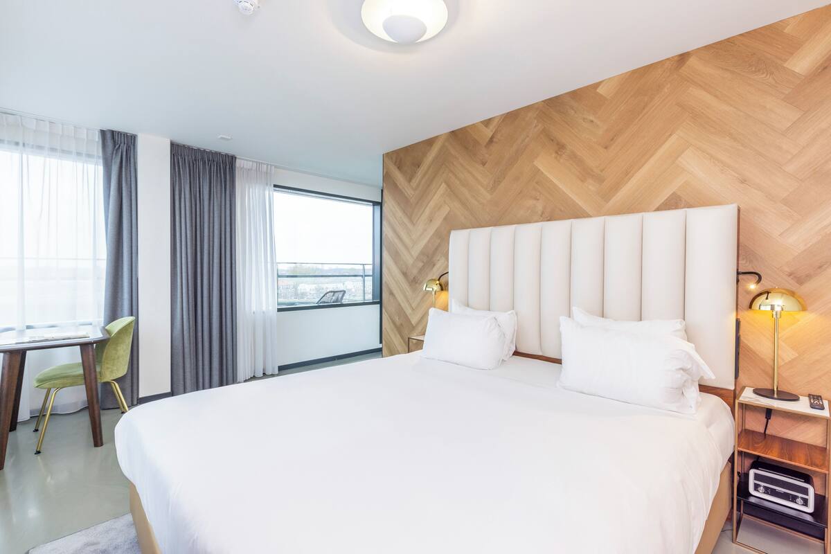 Une chambre d’hôtel avec un grand lit, un bureau avec une chaise, une fenêtre avec des rideaux et un mur en bois.