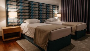 A hotel room with two beds, a large abstract wall art piece above the beds, a wooden nightstand, and a dark curtain on the window. - Mediterranean Villas (Santa Cruz)