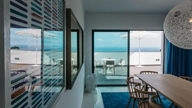 A modern room with a large window overlooking a terrace. There is a dining table with chairs, a blue rug, and a unique spherical light fixture. - Mediterranean Villas (Santa Cruz)