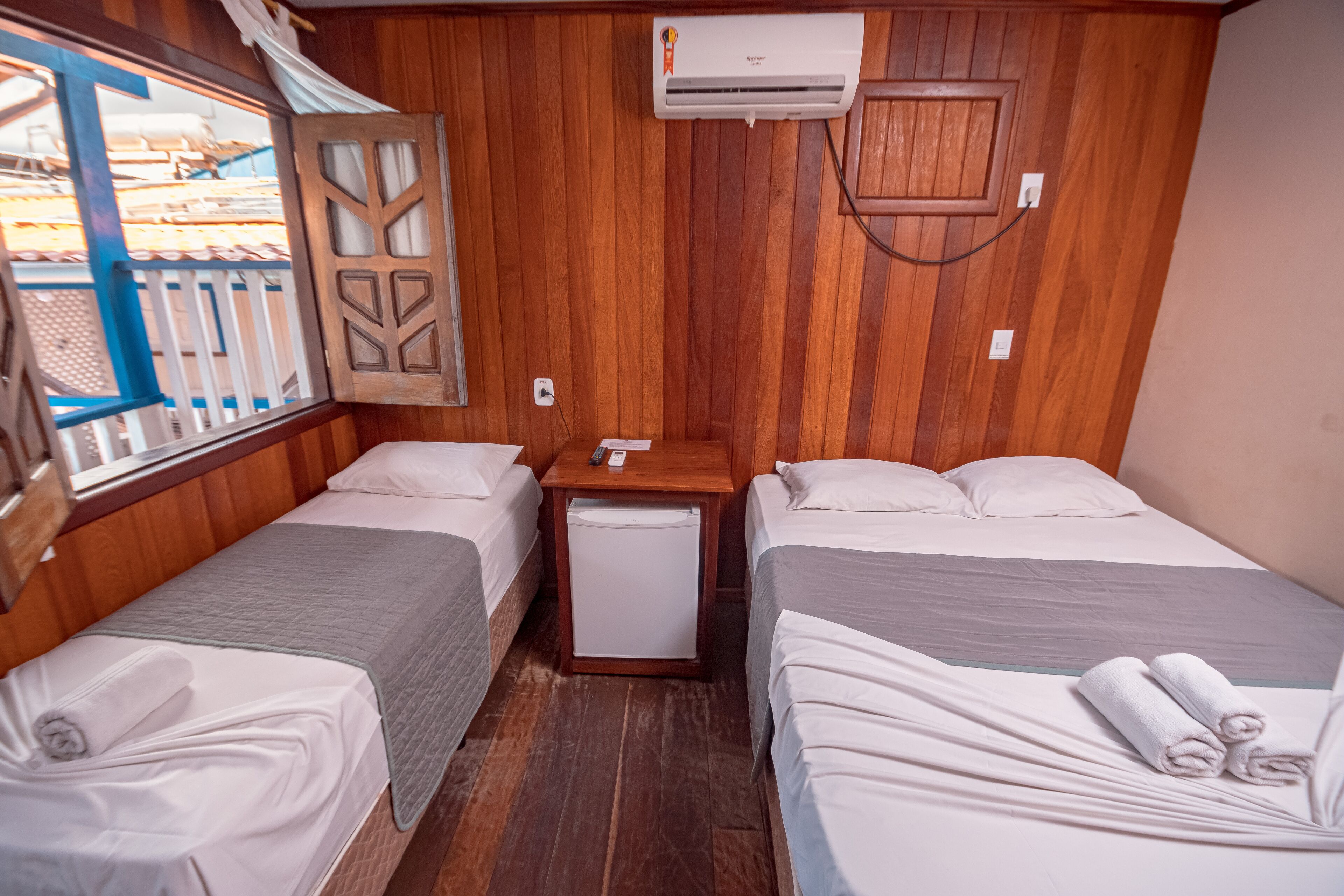 A compact room with two single beds, a small table, a wall-mounted air conditioner, and a window with a view of a wooden deck.
