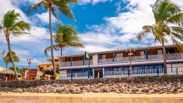 A beachfront hotel with a two-story structure, a stone wall in front, and palm trees.