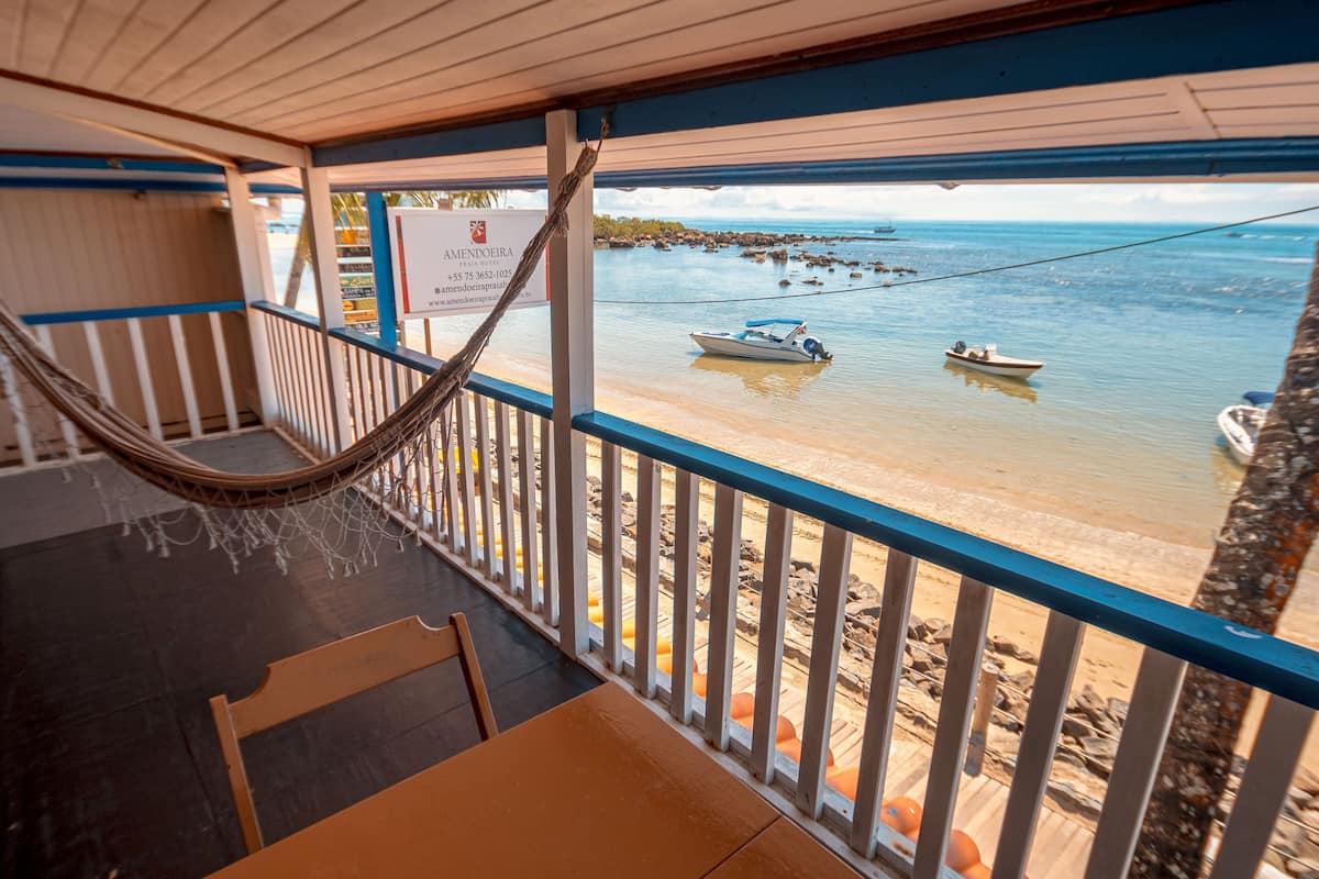 A beachside balcony with a hammock, a wooden table, and a view of boats on the water.