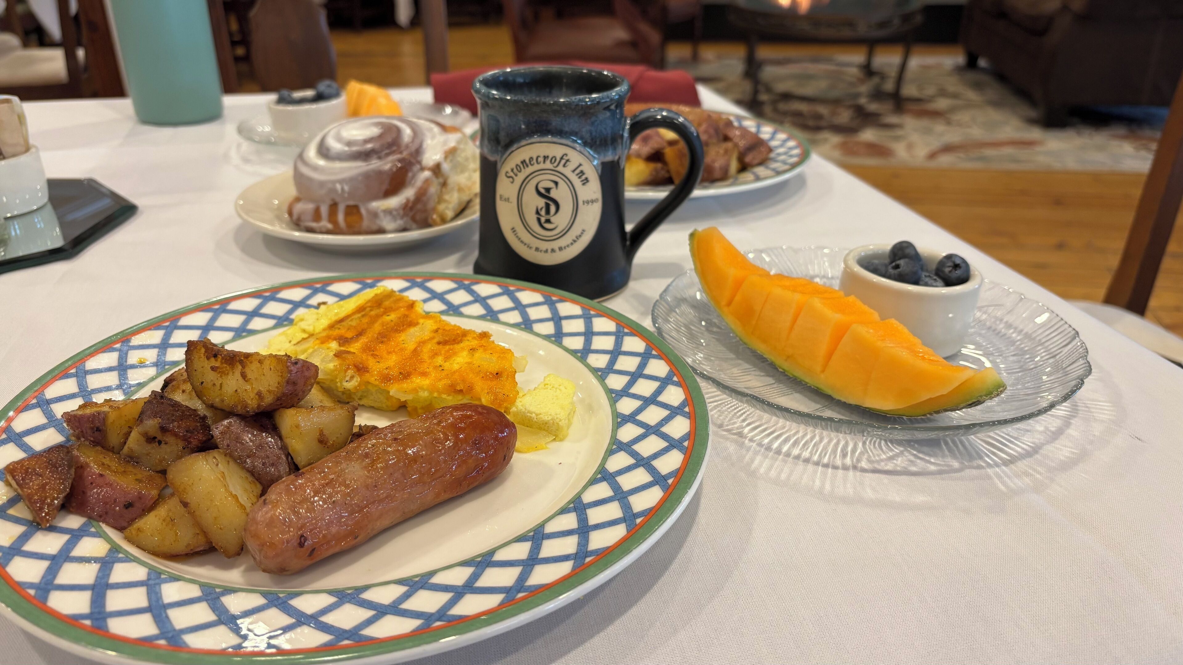 A breakfast plate with sausages, potatoes, and scrambled eggs, a mug, and a plate of fruit.