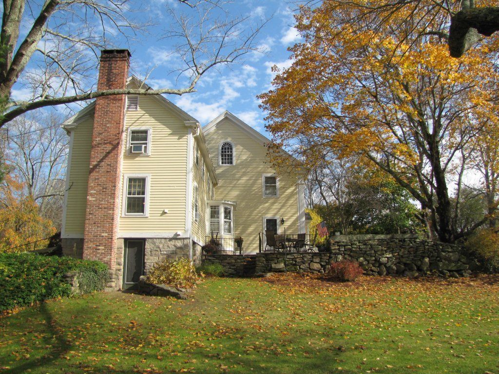 A two-story house with a brick chimney, a stone wall, and a lawn with fallen leaves.