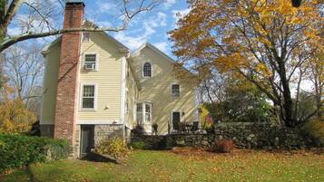 A two-story house with a brick chimney, a stone wall, and a lawn with fallen leaves.