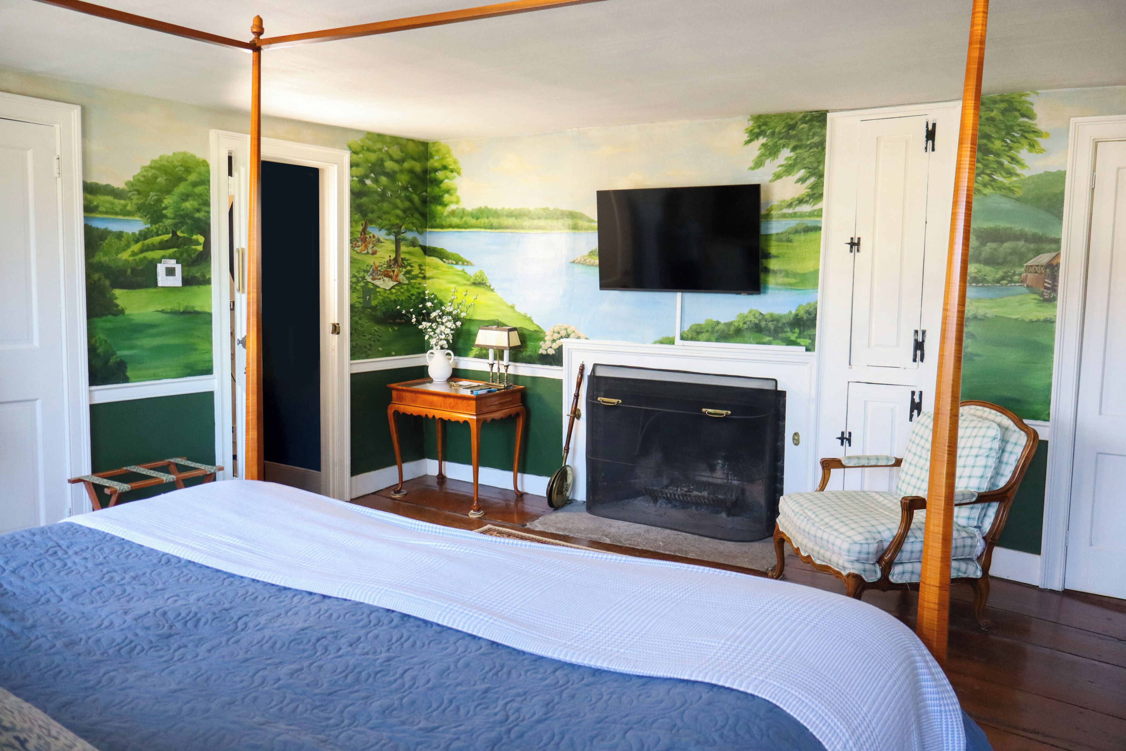 A bedroom with a four-poster bed, a fireplace, a television mounted on the wall, and a scenic mural on the wall.