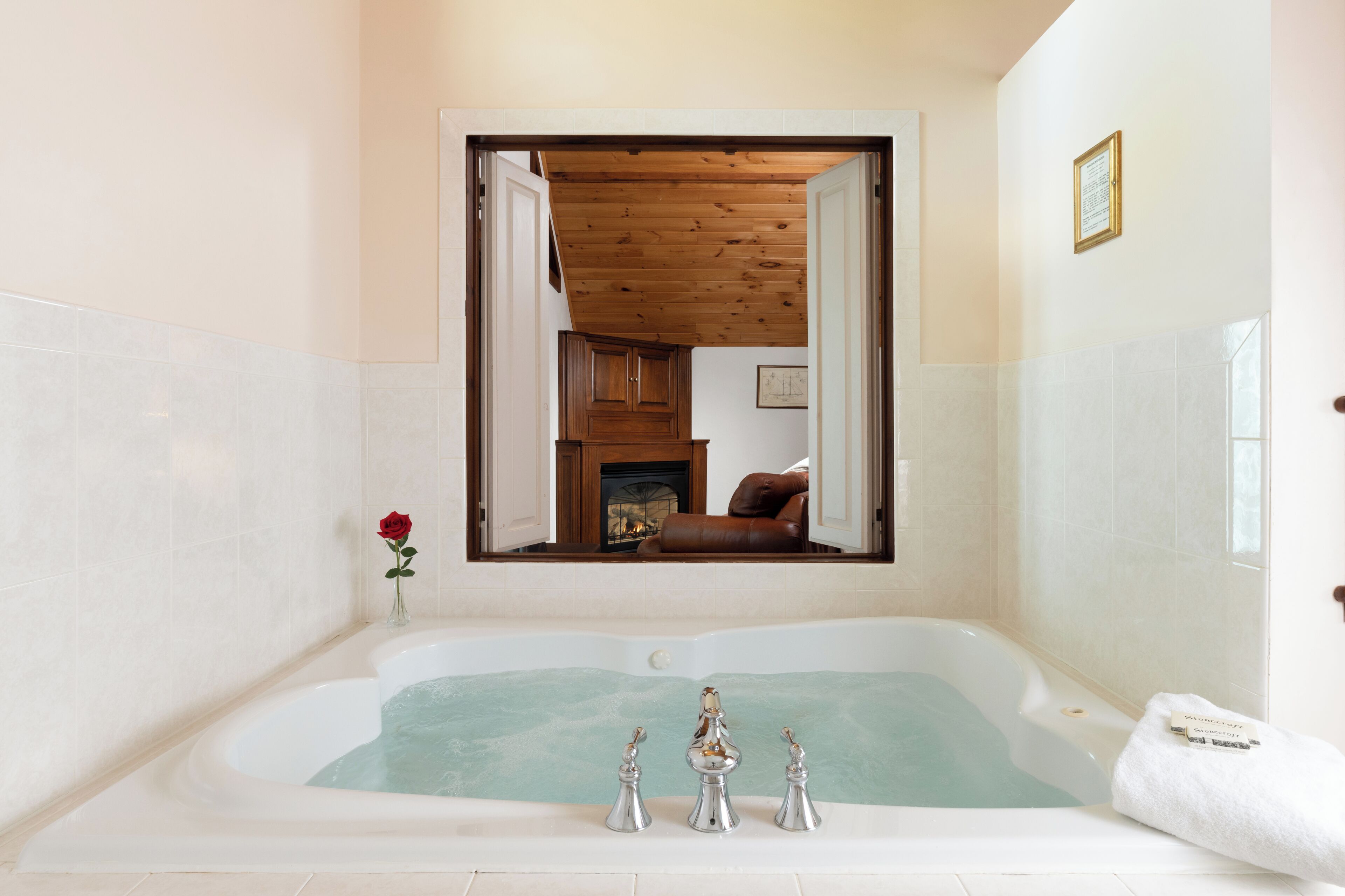 A bathroom with a bathtub, a mirror reflecting a room with a fireplace, and a rose on the wall.