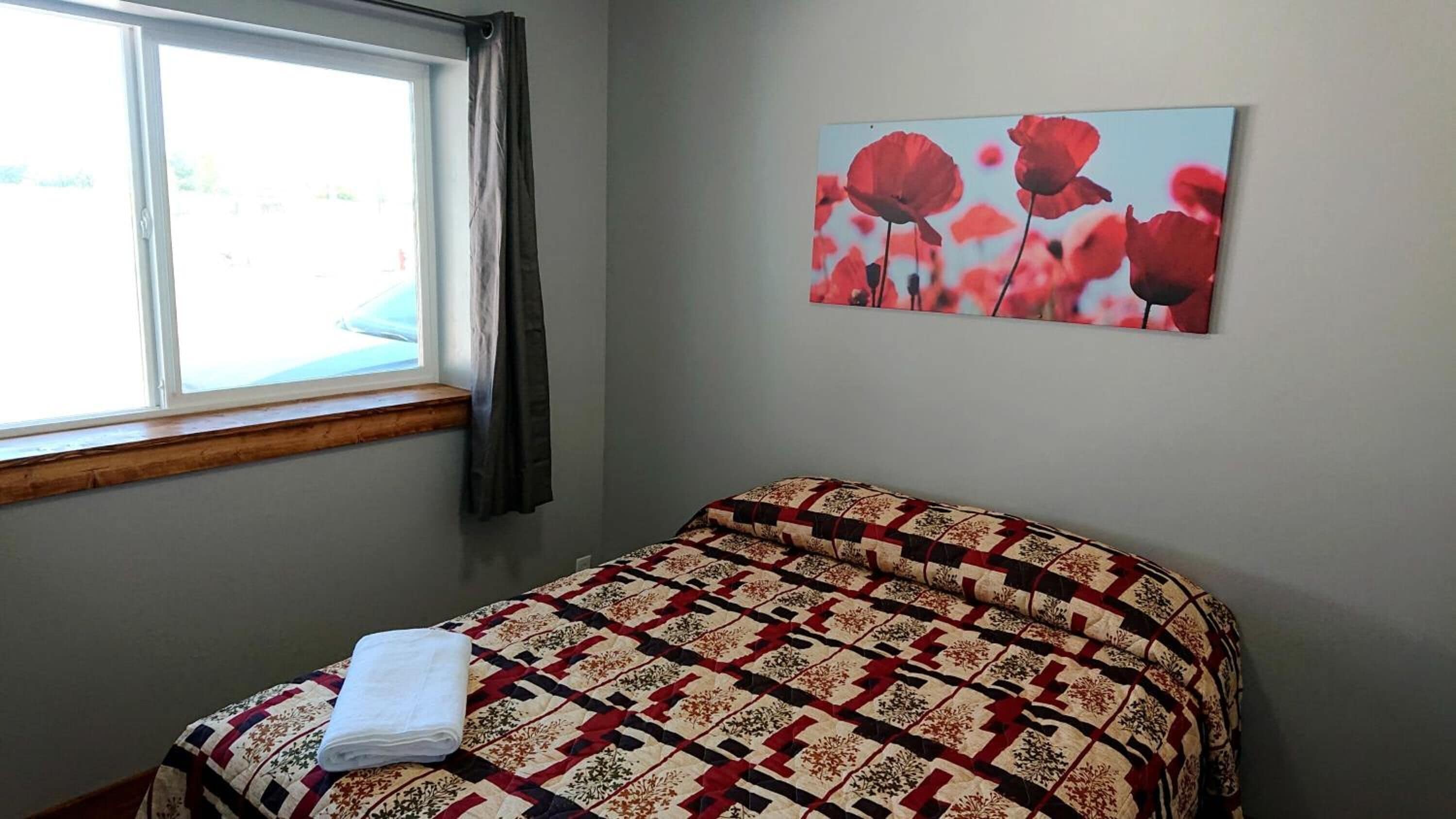 Une chambre à coucher avec un lit, une fenêtre avec des rideaux et un mur orné d’un tableau représentant des coquelicots.