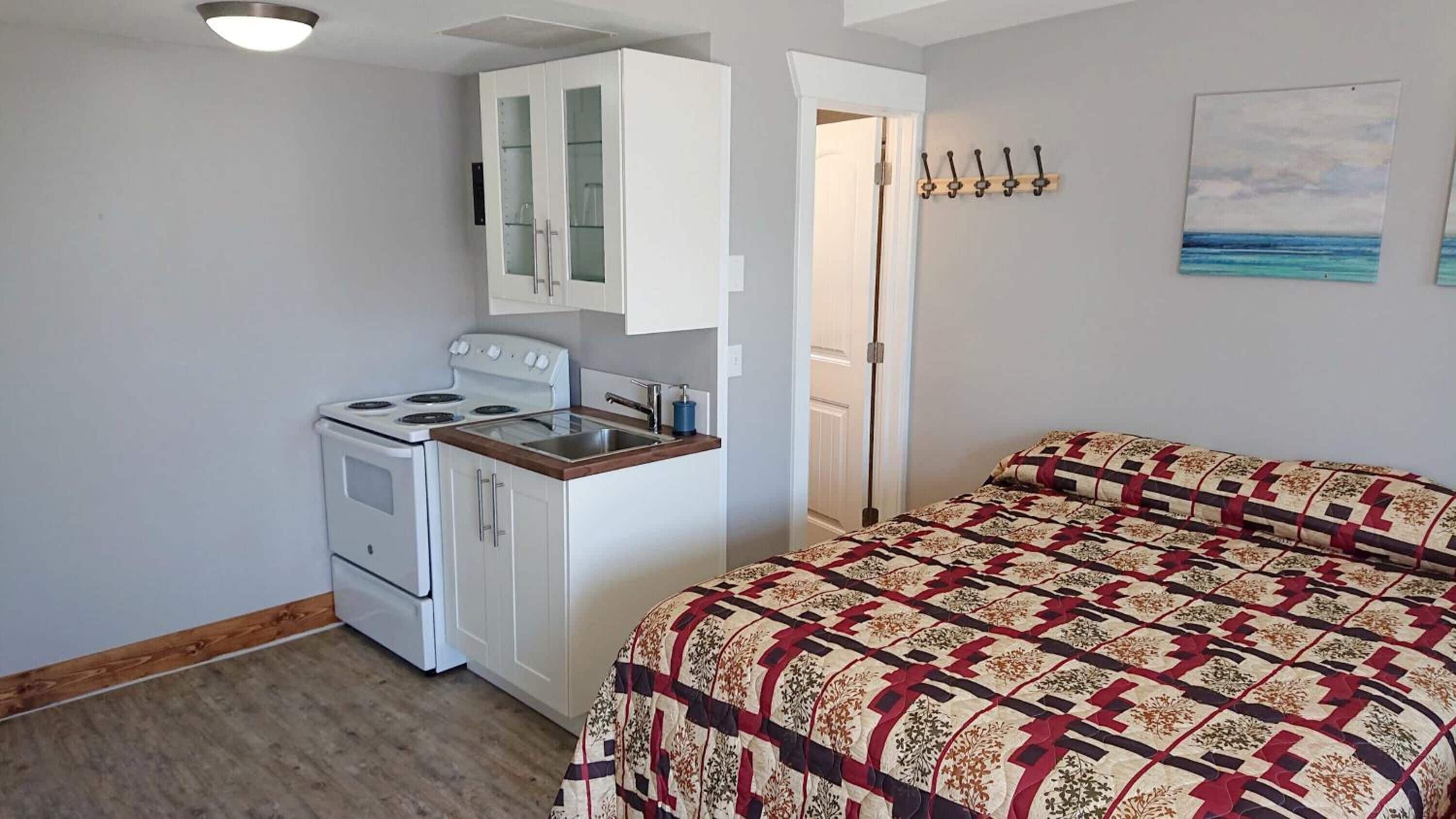 A compact room with a kitchenette, a bed with a patterned quilt, and a door leading to another room.