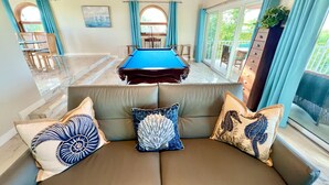 A pool table in a room with a wooden ceiling, a sofa with decorative pillows, and a view of the outdoors through large windows. -  “RENOVATED Gorgeous House with ocean views from balconies, with Hot Tub & Pool” (Marathon)