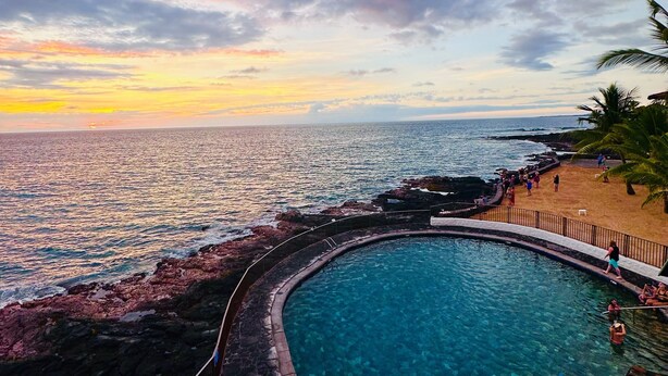 A pool with clear water, surrounded by a stone wall, and overlooking the ocean with a sunset in the background.