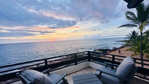 A balcony with two chairs and a small table, overlooking a beach with people and palm trees.