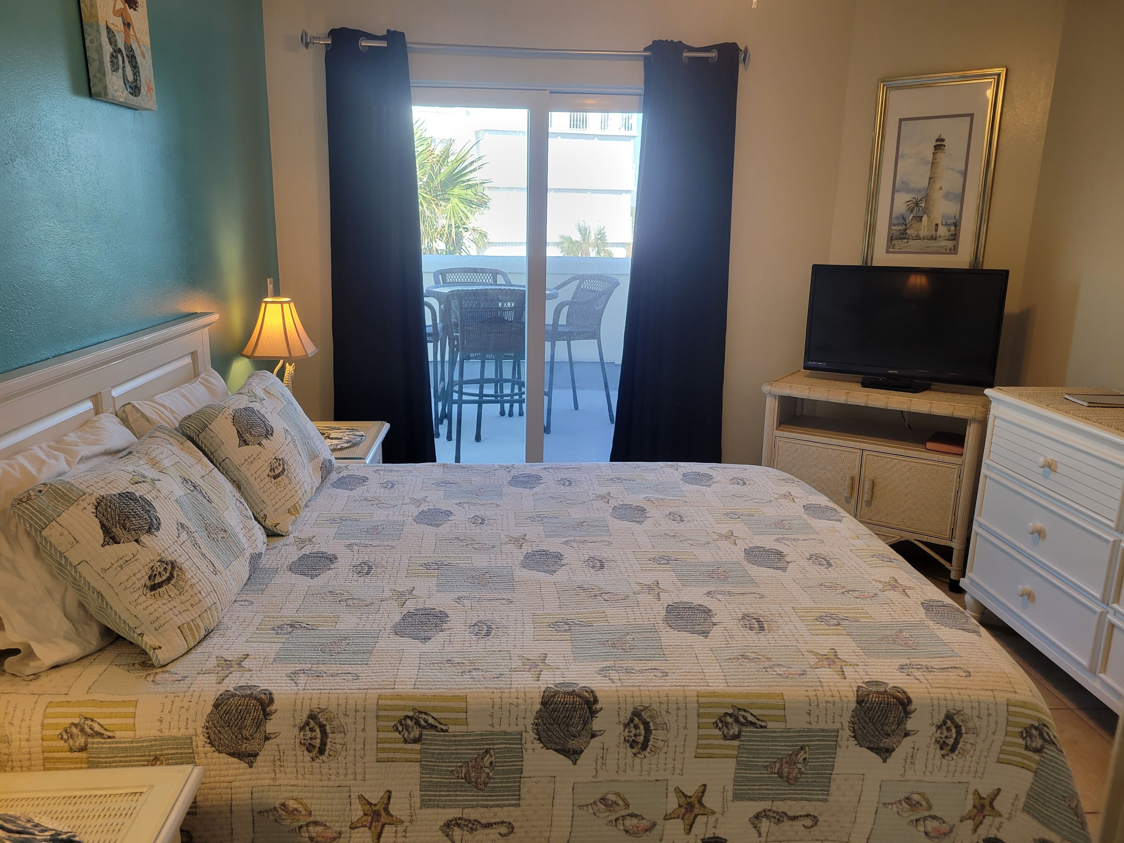 A neatly arranged bedroom with a bed, a television on a stand, a dresser, and a view of the outside balcony.