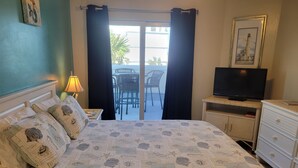 A neatly arranged bedroom with a bed, a television on a stand, a dresser, and a view of the outside balcony.