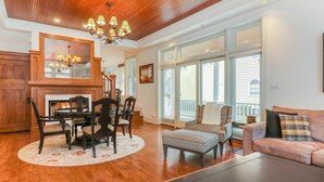 A spacious living room with a wooden ceiling, a fireplace, a dining area with a round table and chairs, and a large window with blinds.