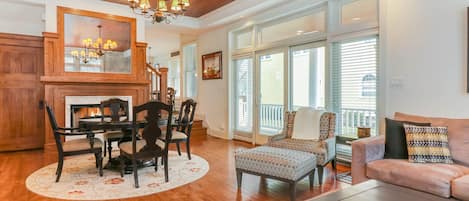 A spacious living room with a wooden ceiling, a fireplace, a dining area with a round table and chairs, and a large window with blinds.