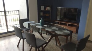 A modern living room with a glass-top dining table surrounded by four chairs, a large flat-screen TV, and a bookshelf.