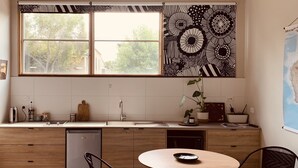 A modern kitchen with a large window, wooden cabinets, a sink, and a dining area with two chairs. - Inner city garden studio in the heart of Footscray (Footscray)