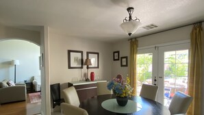 A dining area with a round table, six chairs, and a vase of flowers. There is a kitchen in the background with a window and a door leading outside.