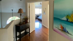A hallway with wooden flooring, a ceiling vent, a lamp, and a painting on the wall.