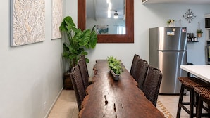 A dining area with a wooden table, chairs, a hanging light fixture, and a kitchen in the background.