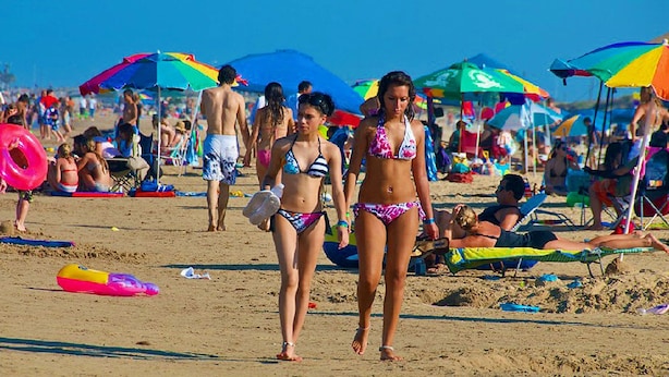A crowded beach with people walking and sunbathing under colorful umbrellas.