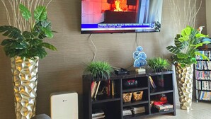A living room with a flat-screen TV mounted on the wall, a bookshelf, and two large potted plants.