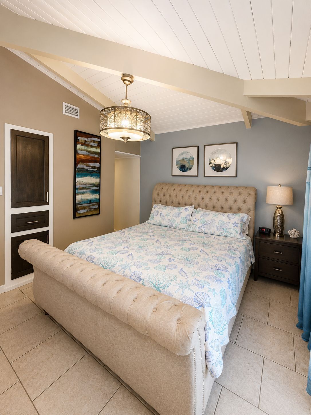 A bedroom with a large bed, a chest of drawers, a bedside table, a lamp, and two wall-mounted pictures.
