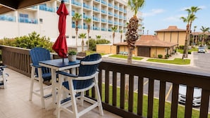 A covered outdoor seating area with a table and chairs, a red umbrella, and a view of a hotel building and palm trees.