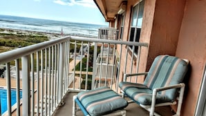 A balcony with two striped chairs and a small table, overlooking a swimming pool and a view of the ocean.