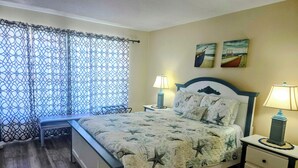 A bedroom with a bed, two bedside tables, a bench, a window with a patterned curtain, and two wall-mounted paintings.