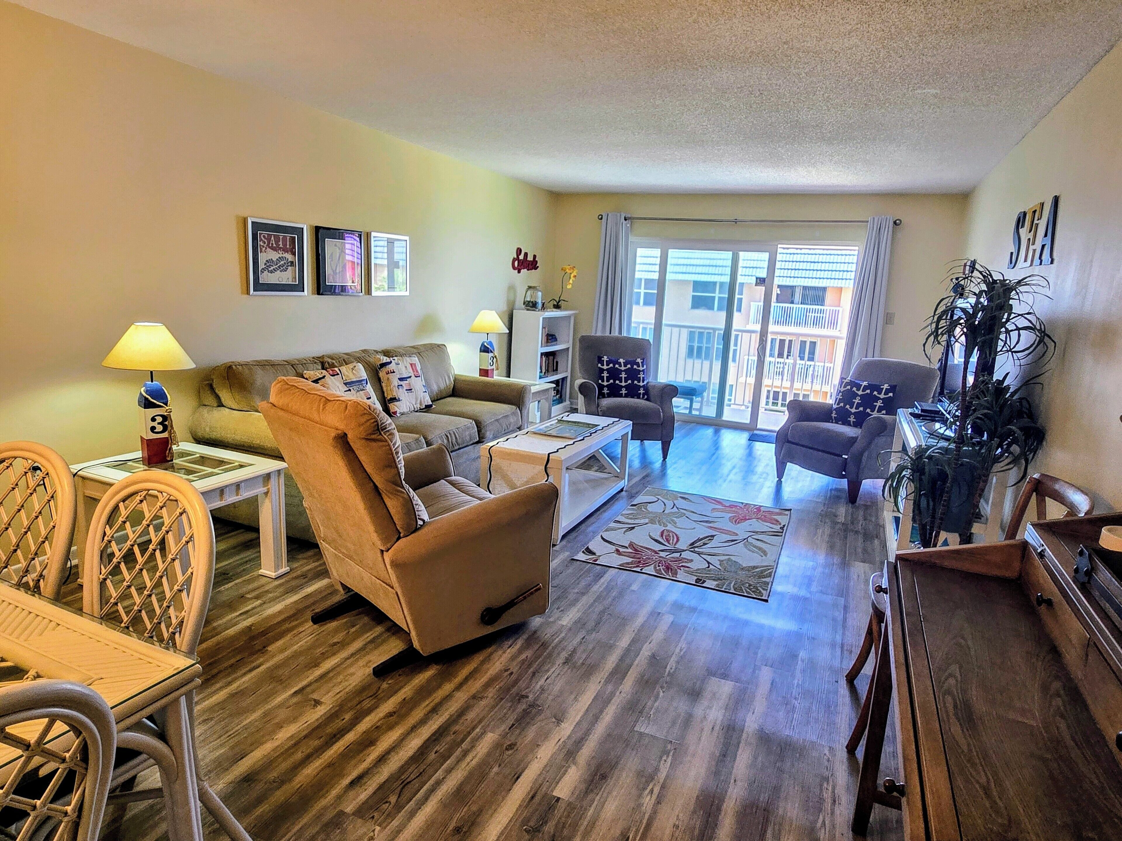 A living room with a sofa, armchairs, a coffee table, and a dining area with chairs. There is a large window with curtains, a plant, and a view of a building outside.