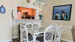 A dining area with a glass-top table surrounded by white wicker chairs, a kitchen in the background, a star decoration, and a framed picture of pelicans.