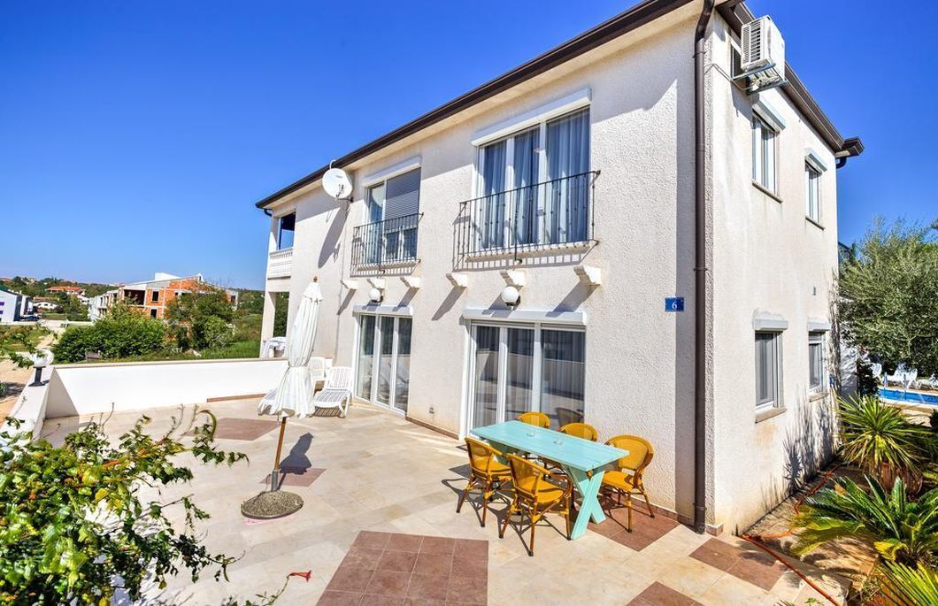A two-story building with a patio area featuring a table and chairs, a pool, and a garden.