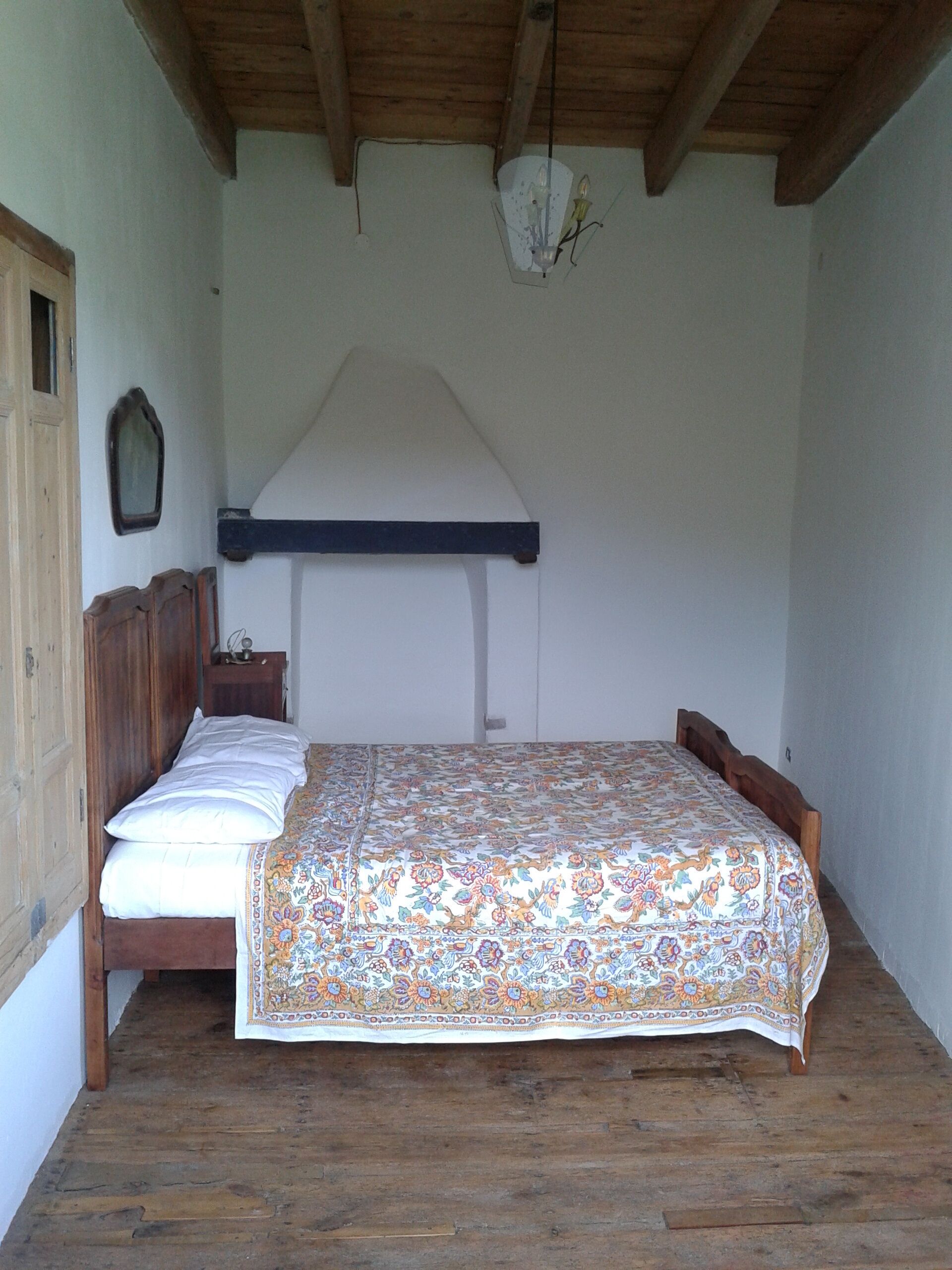 A bedroom with a wooden ceiling, a bed with a patterned bedspread, a nightstand, and a wall-mounted light fixture.