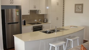 A modern kitchen with white cabinets, a stainless steel refrigerator, a double sink, and a small island with stools. - C and C Tanunda (Tanunda)