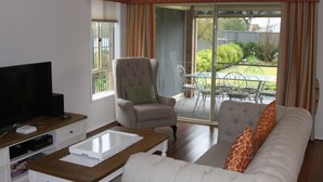 A living room with a sofa, armchair, and coffee table. There is a television on a stand and a window with curtains. - C and C Tanunda (Tanunda)