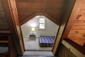 Une petite chambre mansardée avec un plafond en pente, une petite table de chevet avec une lampe, un lit recouvert d’une couverture à carreaux et une fenêtre laissant entrer la lumière naturelle.