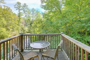 Une terrasse en bois avec une table ronde et deux chaises en osier, entourée d’une végétation luxuriante et d’arbres.