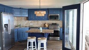 A modern kitchen with blue cabinets, a stainless steel refrigerator, a built-in oven, and a large island with stools. - On The Bay In Sea Isle: Fishing, Family, Fun! (Galveston)