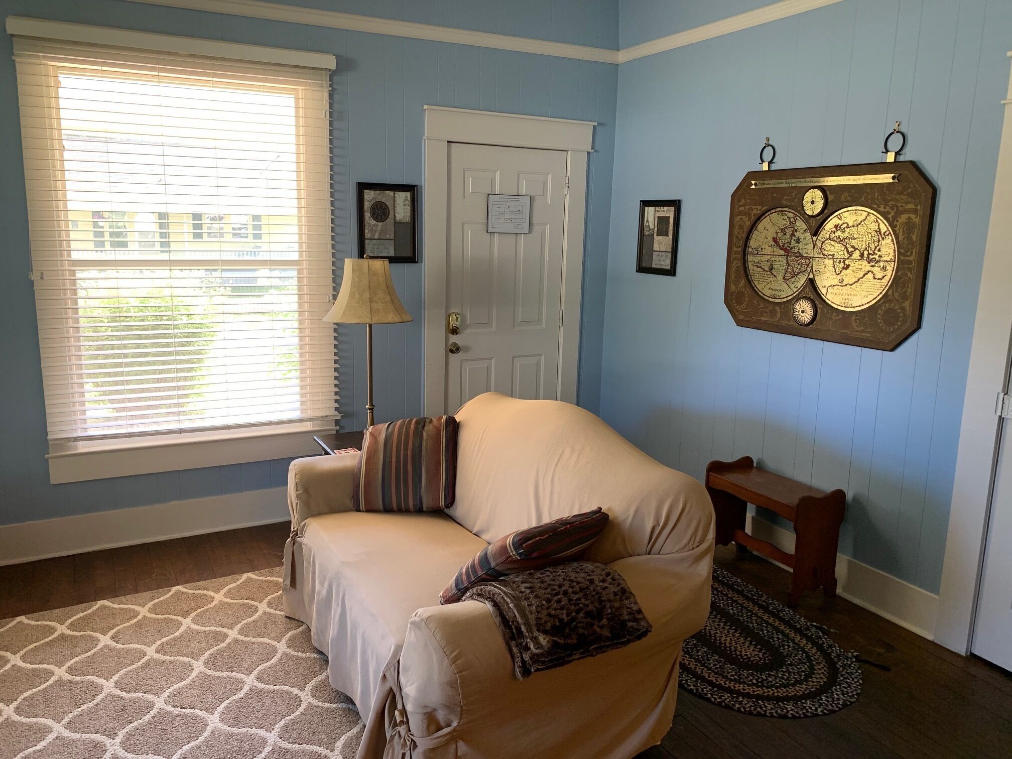 Una habitación con un sofá color crema, una mesita de madera, un adorno de globo en la pared, una puerta y una ventana con persianas.