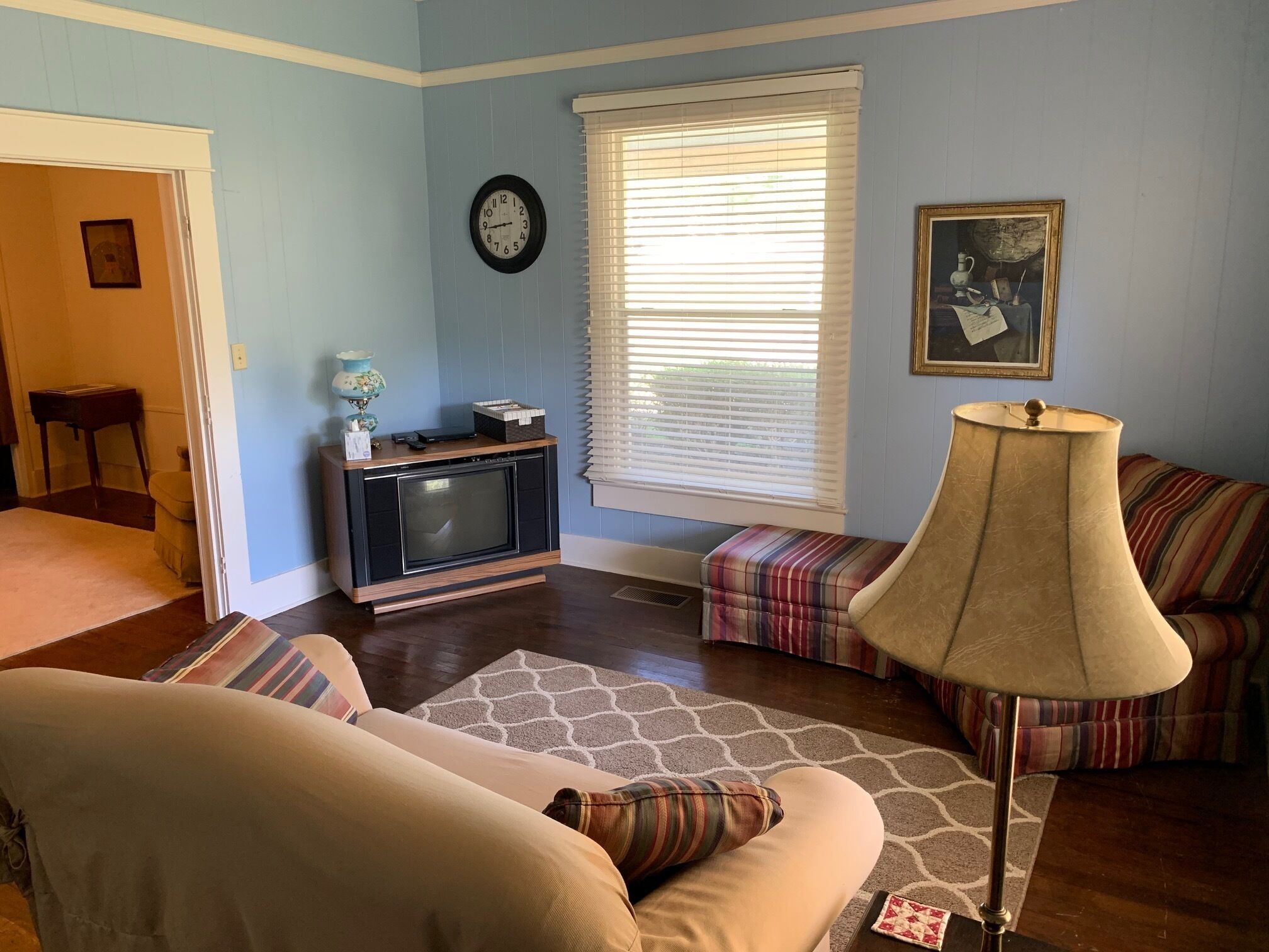 Uma sala de estar com televisão, luminária, sofá, janela com persianas e um quadro na parede.