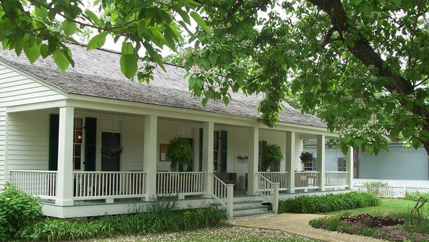 Une maison à deux étages avec un porche couvert, une cour avant avec une pelouse et des parterres de fleurs, et une clôture blanche en bois.