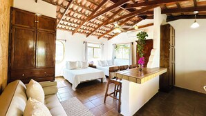 A cozy room with a wooden ceiling, a bed with white bedding, a sofa with cushions, a small table with a vase of flowers, and a window with curtains. - CABO BOHEMIA Tropical Garden Studios (Cabo San Lucas)