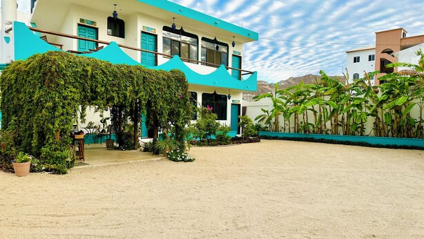 A two-story building with a covered outdoor area, surrounded by greenery and a sandy courtyard. - CABO BOHEMIA Tropical Garden Studios (Cabo San Lucas)