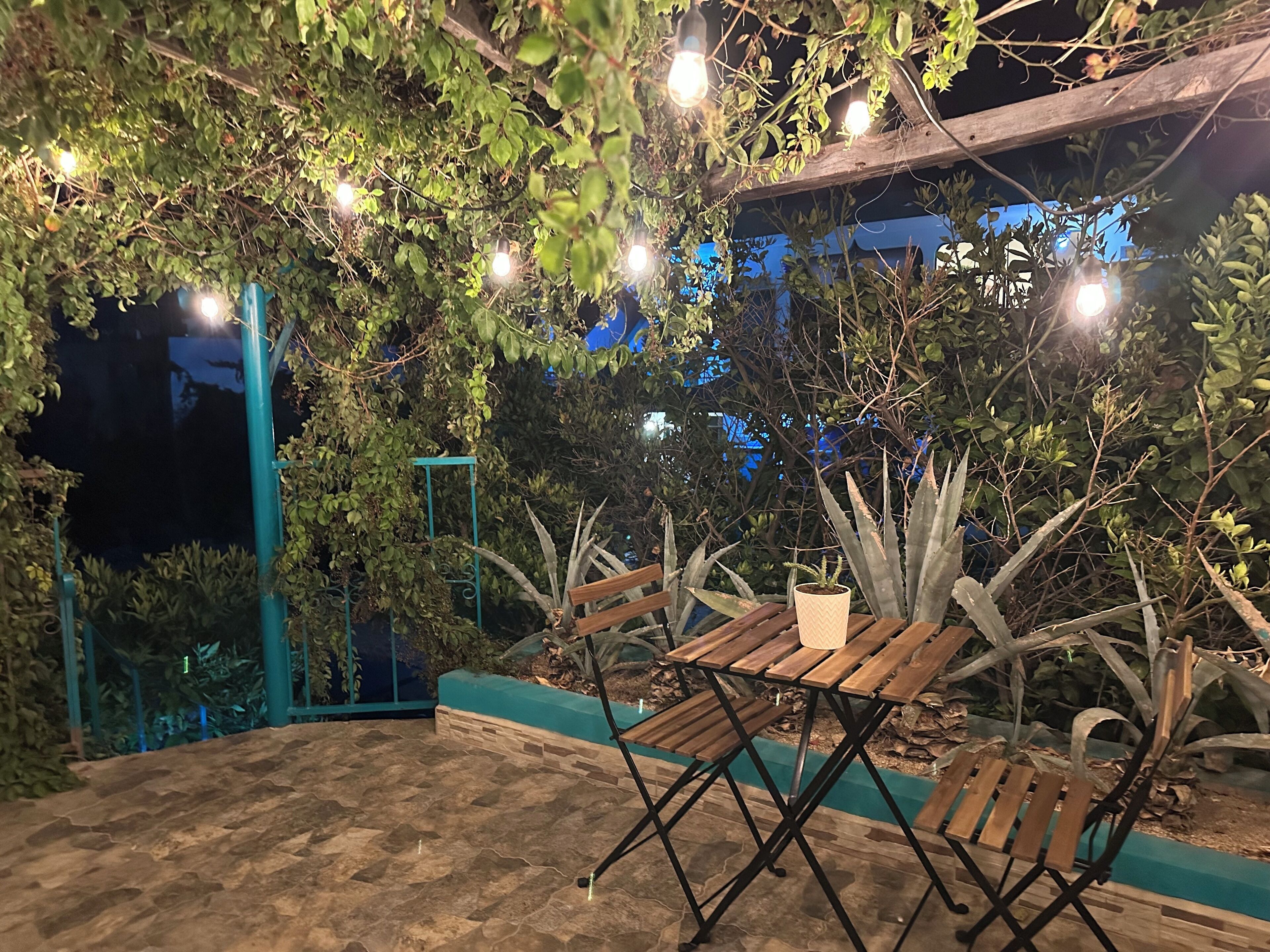 Un espacio patio al aire libre con una mesa y sillas de madera, rodeado de follaje y luces colgantes.