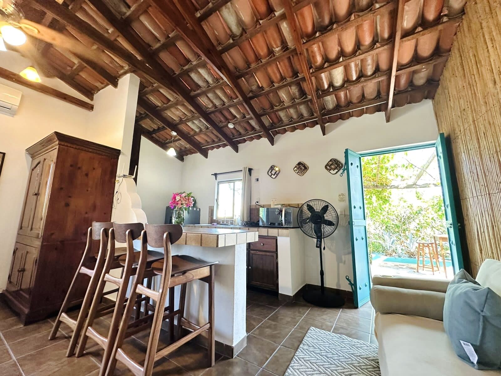 A cozy kitchen with a wooden ceiling, a dining area with wooden chairs, a small island with a sink, and a fan near the window.