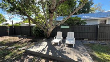 Ein Garten mit einer Holzterrasse, zwei weißen Stühlen, einem Baum und einem Zaun.